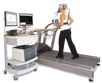 Photo of a woman walking on a treadmill while wearing a clear mask over her nose and mouth, and attached to a cart holding medical equipment.